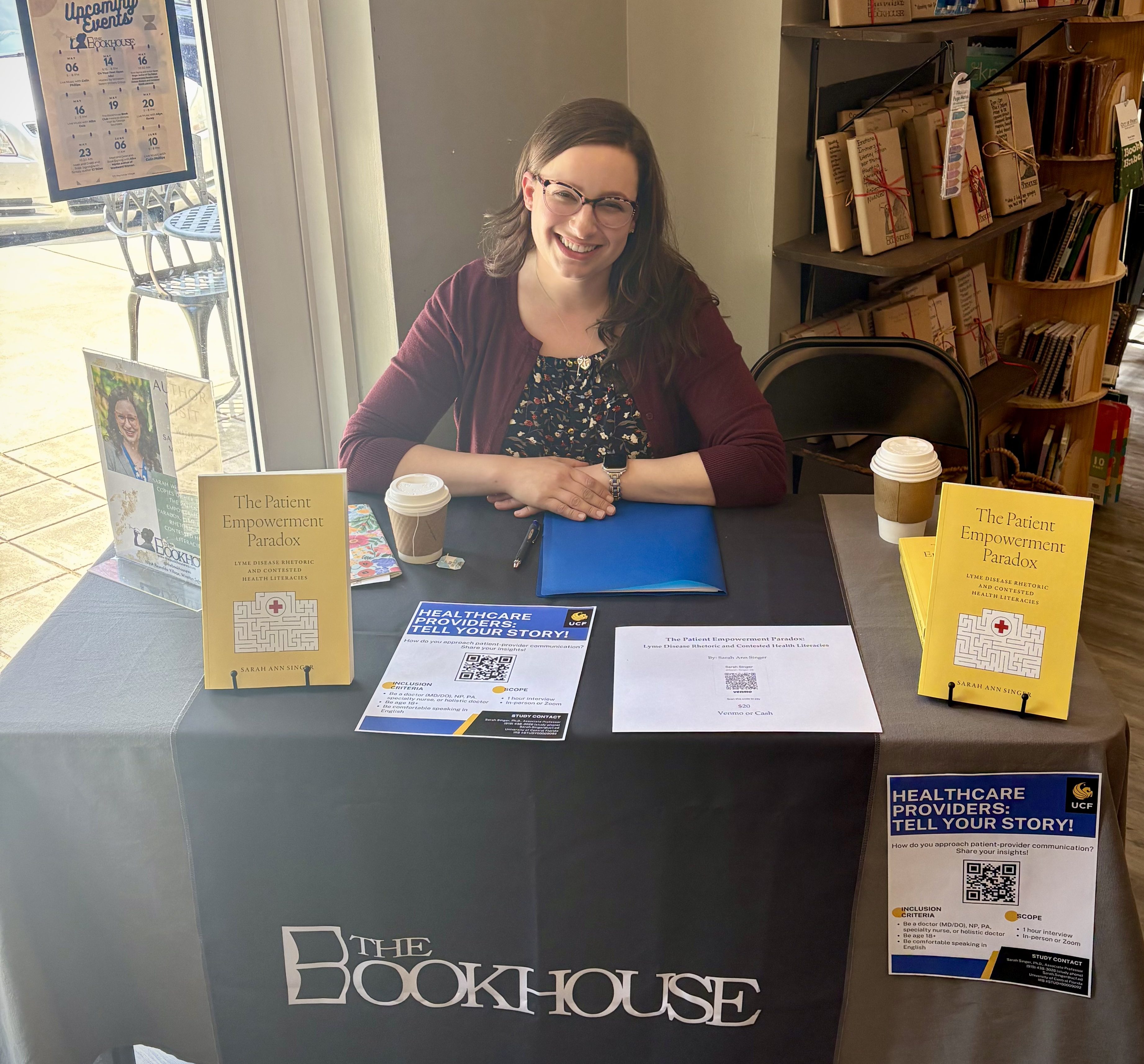 Sarah Singer, a white woman with glasses and brown hair, sits behind a table and between two copies of her book, The Patient Empowerment Paradox. A bookshelf is visible in the background.